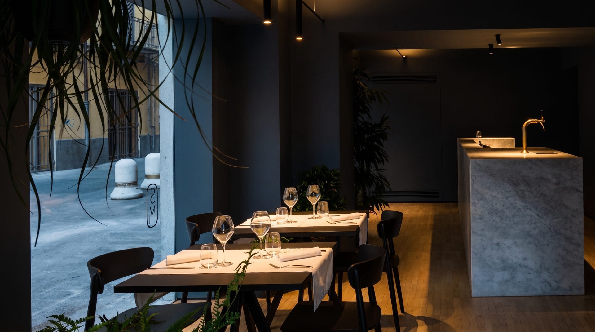 La sala di Sobremesa a cena: tavoli apparecchiati con tovaglioli di lino sotto i faretti, il bancone di marmo e la piazza oltre la vetrata
